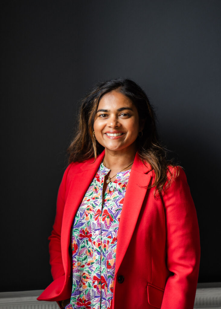 A portrait picture of Devina. She is wearing a red jacket and a floral shirt. She has shoulder length hair.