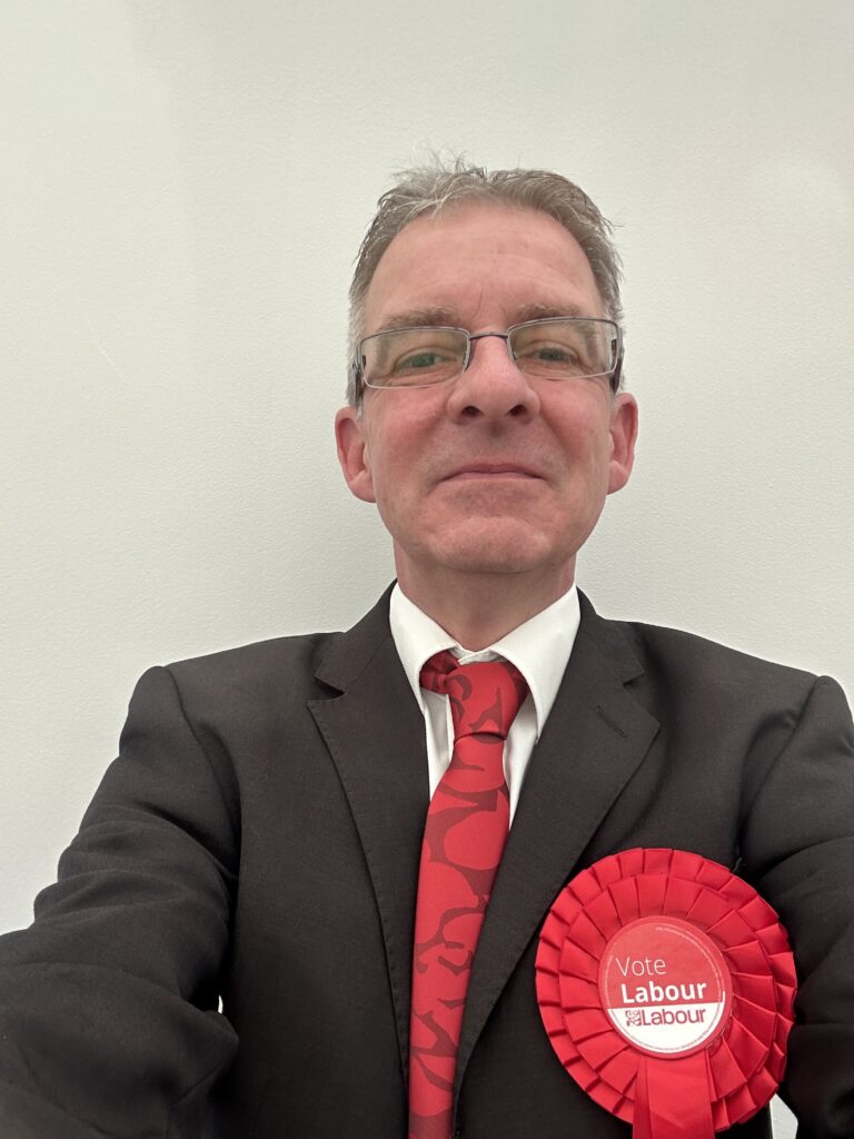 A picture of Geoff Budd a Man wearing a suit jacket with a white shirt and red Tie. He is also wearing a Red Rosette with the word Vote Labour.